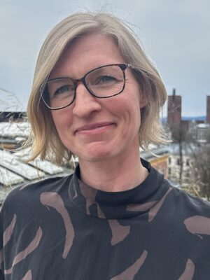 Portrett av kvinne med kort, blondt hår med bylandskap i bakgrunnen. Foto.