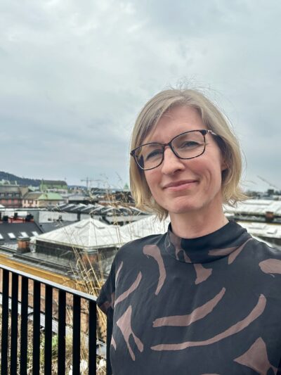 Portrett av lyshåret kvinne med briller med bylandskap i bakgrunnen