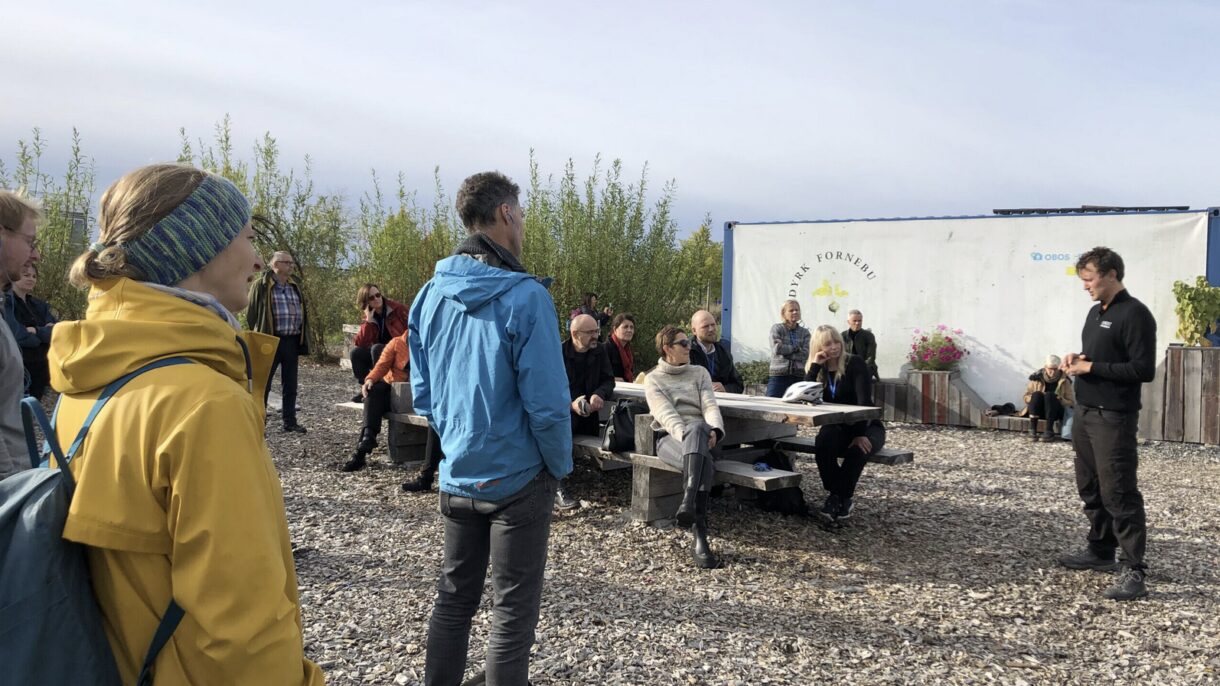 En gruppe mennesker får omvisning utendørs. Noen sitter og noen står. Foto.