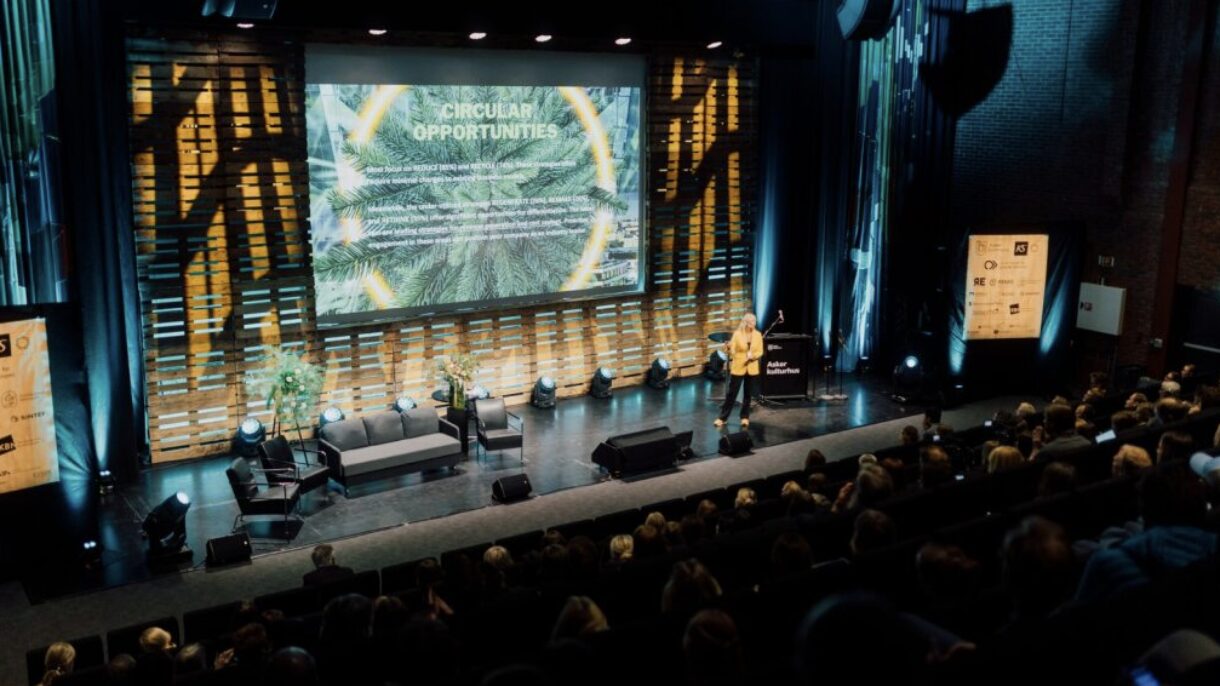 Konferanse med kvinne kledd i gul dressjakke på scenen. I bakgrunnen slide med teksten Circular Oppurtunites. Foto.