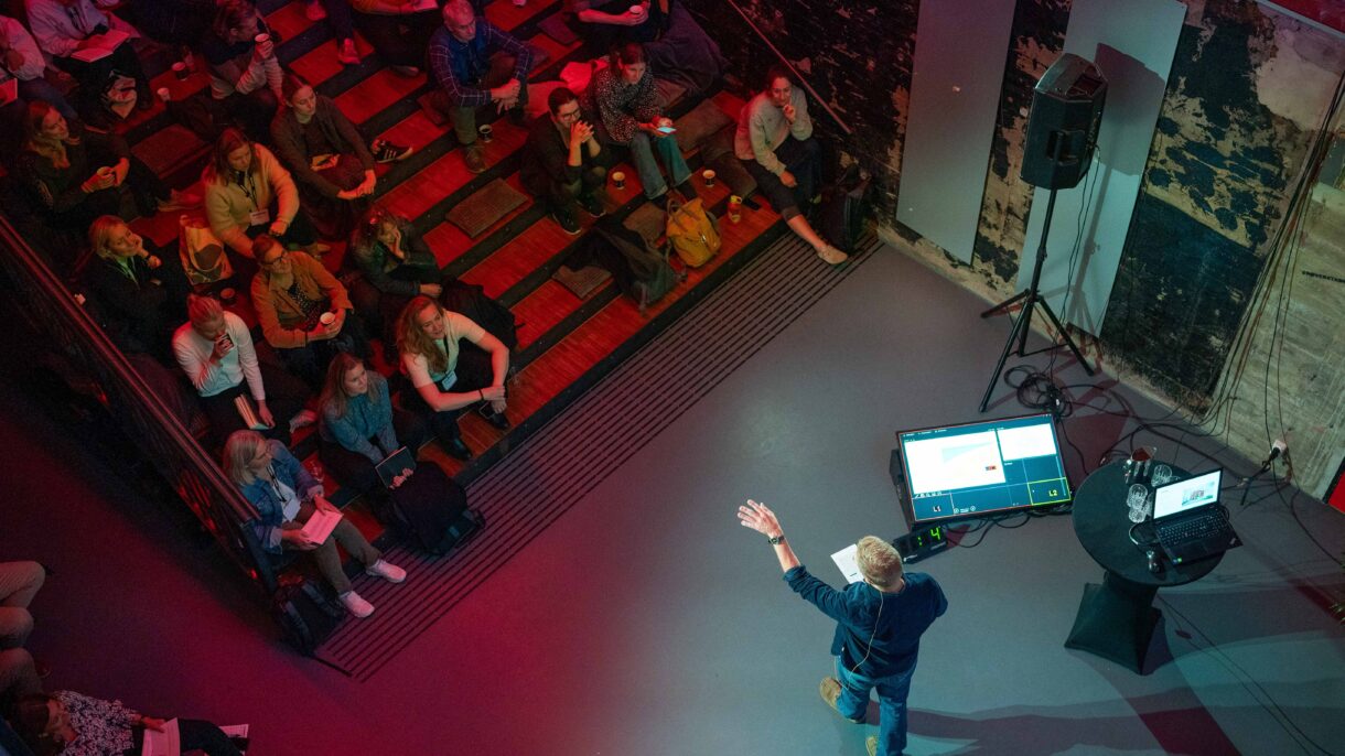 Mann holder foredrag for publikum som sitter i trapper. Foto.