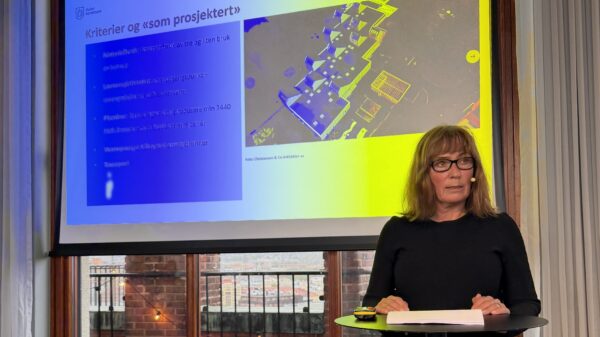 Kvinne holder foredrag. I bakgrunnen en slide med teksten "Kriterier og "som prosjektert" sammen med et bilde av en barnehage i fugleperspektiv. Foto.