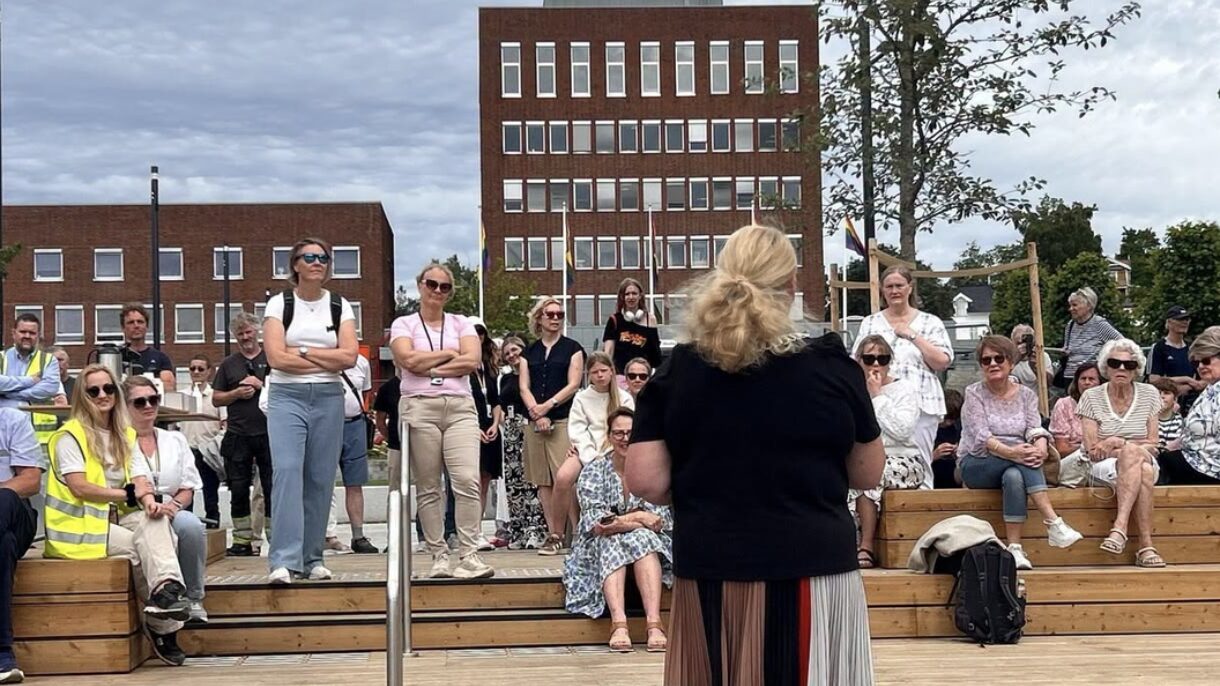 Kvinne holder tale foran publikum utendørs. Foto.