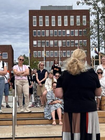 Kvinne holder tale foran publikum utendørs. Foto.