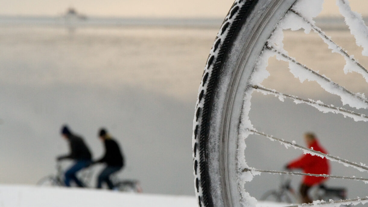 Sykkelhjul med snø hvor vi ser en person kledd i rødt mellom eikene. To personer sykler til venstre i bildet. Foto.