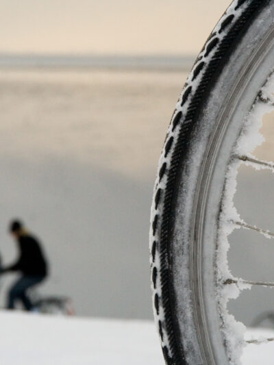 Sykkelhjul med snø hvor vi ser en person kledd i rødt mellom eikene. To personer sykler til venstre i bildet. Foto.