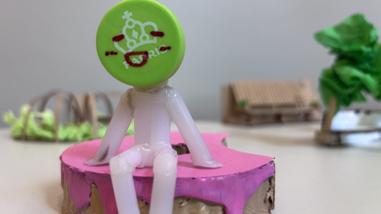 Møbelmodell med figur med farriskork som hode som sitter på rosa benk. Foto.
