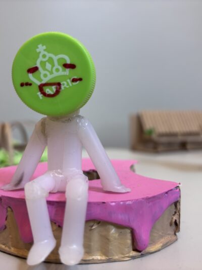 Møbelmodell med figur med farriskork som hode som sitter på rosa benk. Foto.