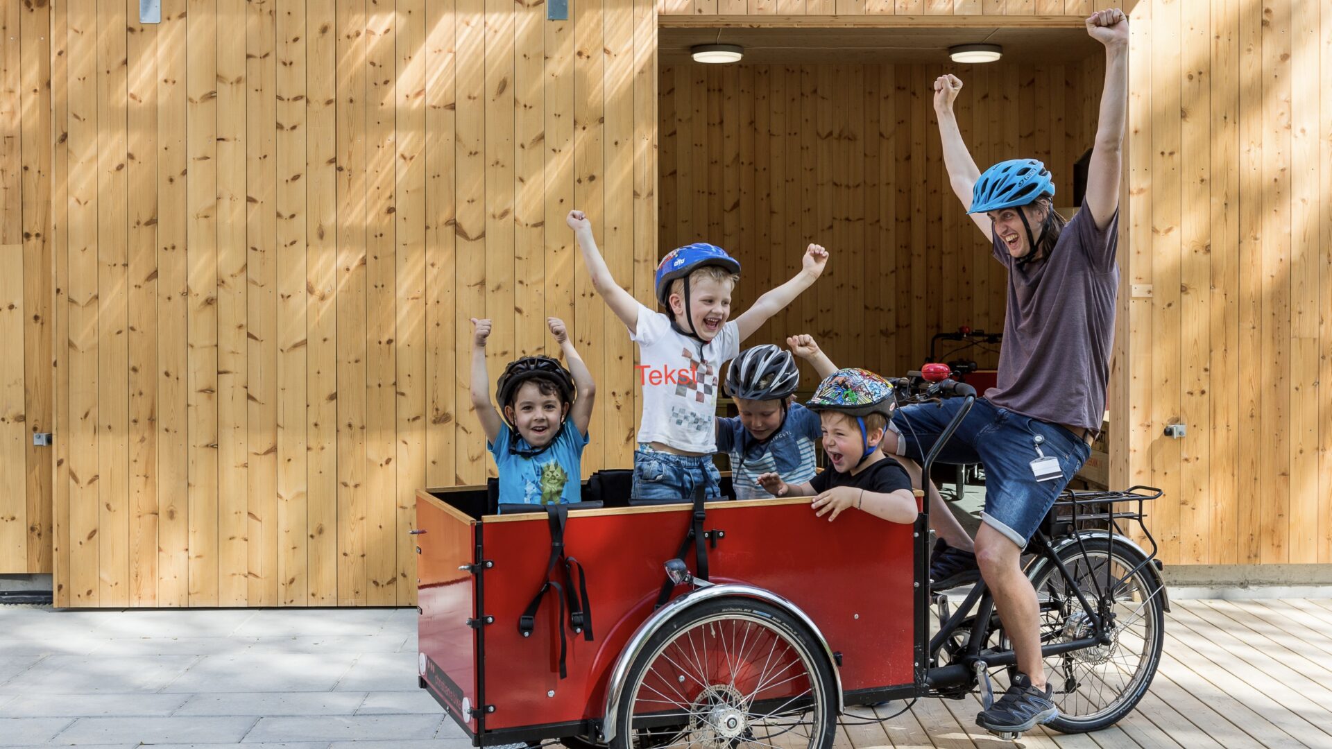 Lastesykkel med fire jublende barn og en voksen som alle jubler og strekker armene i været. Foto.