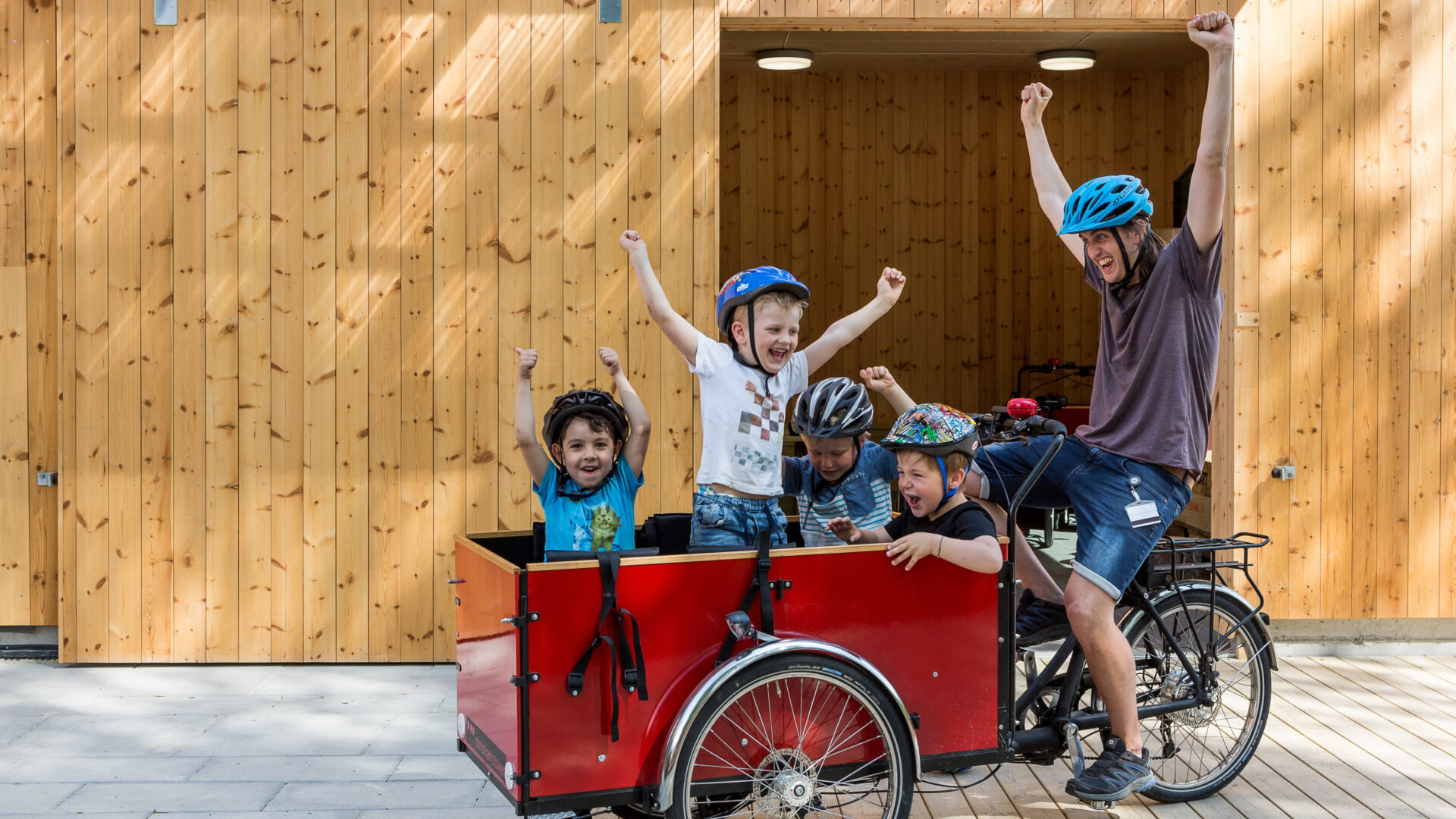 Mann med hendene i været på lastesykkel med fire barn i kasse foran sykkel. Fotografi.