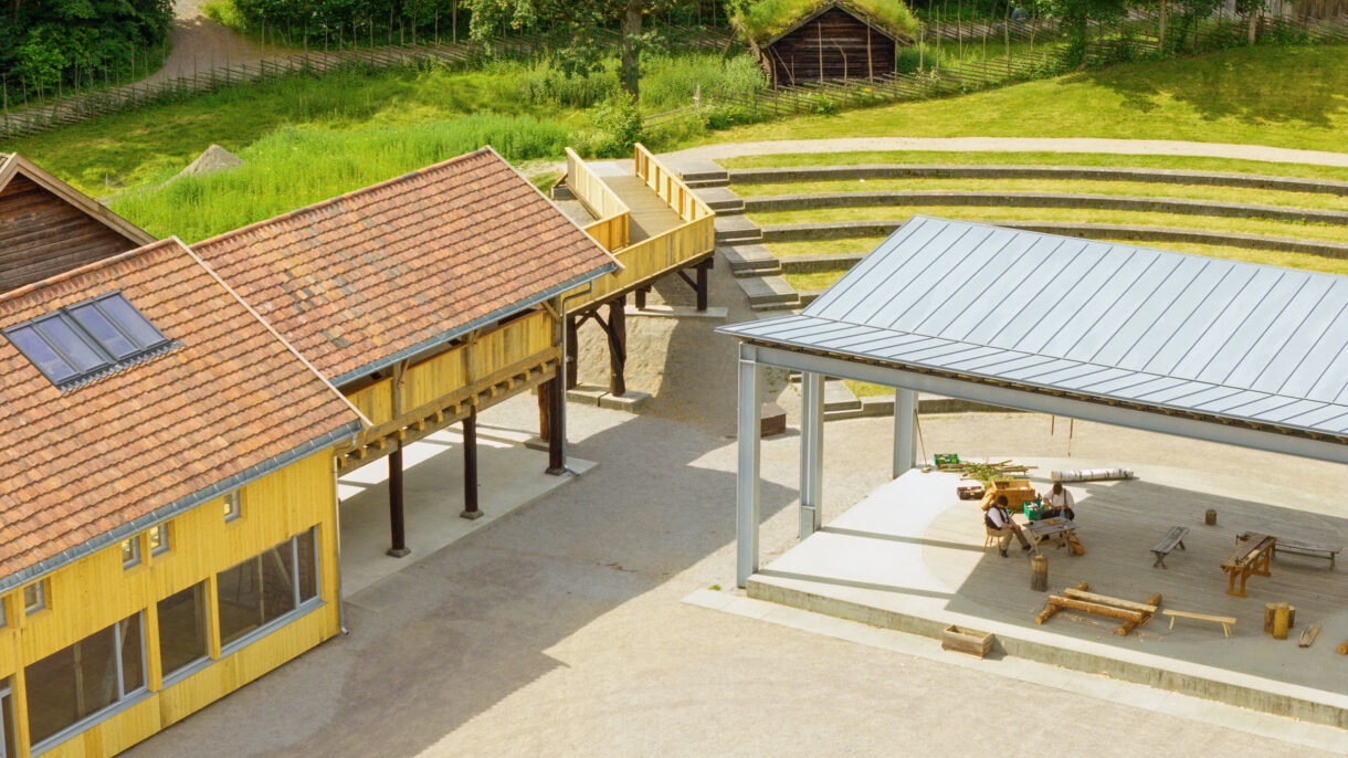 Trehus med rød takstein og plass med overbygg ved større plass. Foto.