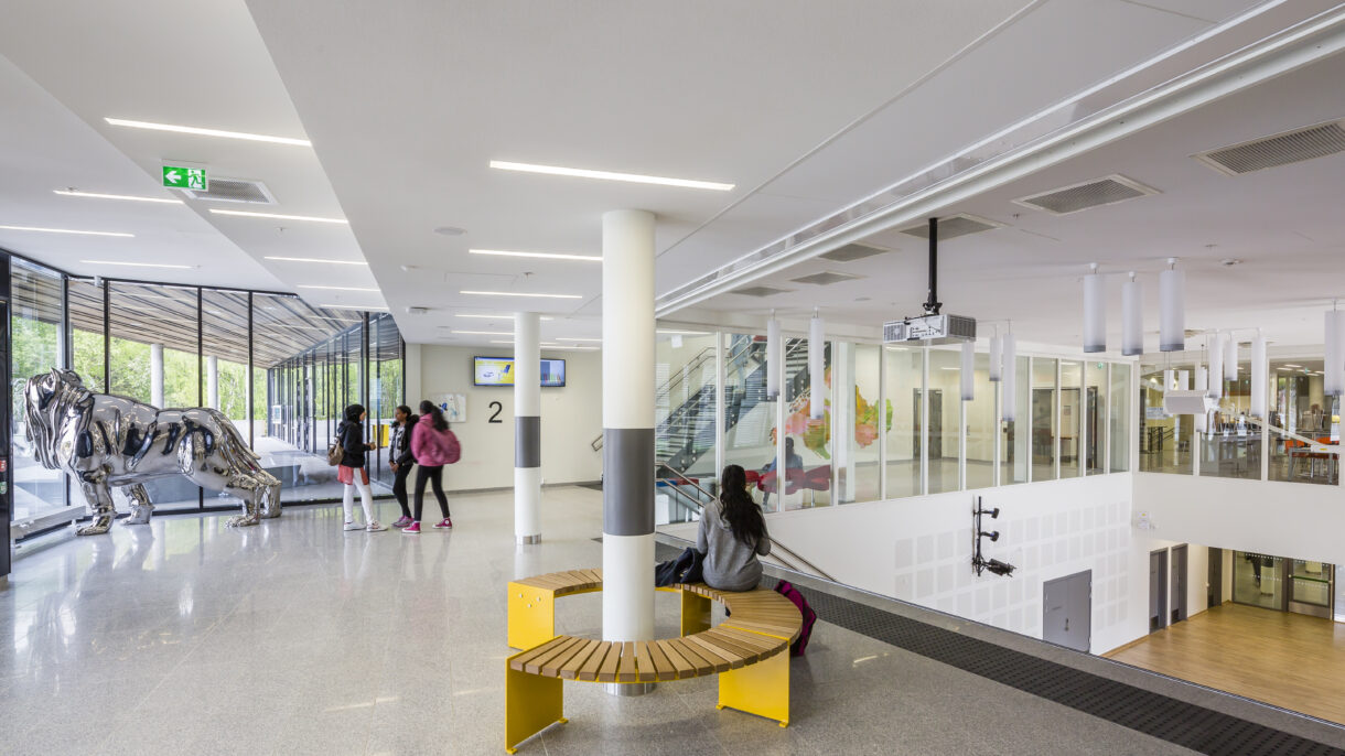 Interiør skole med barn, utsikt til atriumsrom og skulptur.