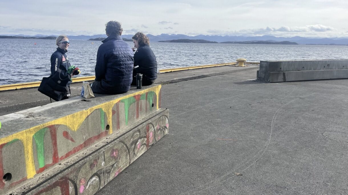 To personer sitter på bærebjelker i betong ved en kaikant og prater med en tredje person som står. Foto.