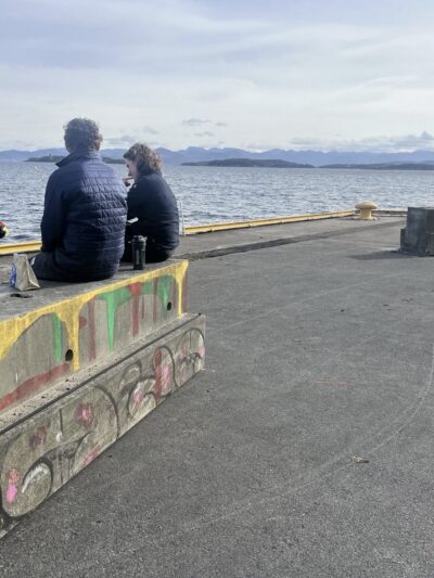 To personer sitter på bærebjelker i betong ved en kaikant og prater med en tredje person som står. Foto.