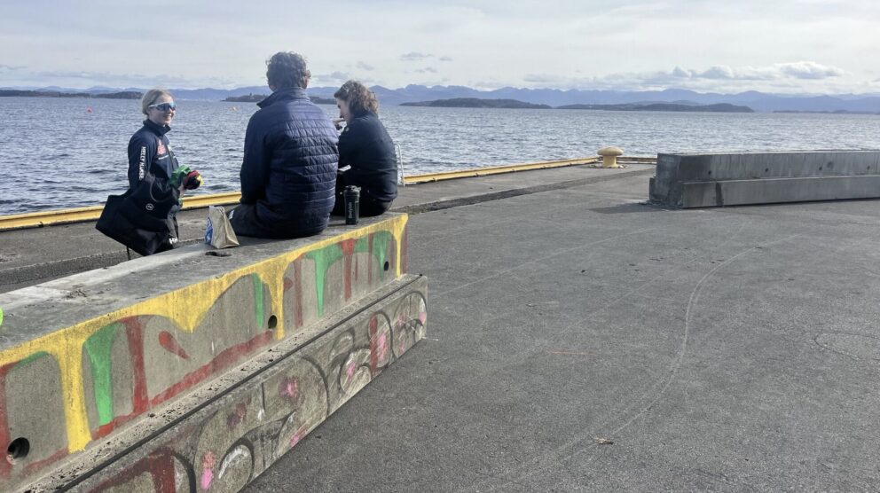 To personer sitter på bærebjelker i betong ved en kaikant og prater med en tredje person som står. Foto.