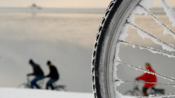 Sykkelhjul hvor man mellom eikene ser en person kledd i rødt som sykler. Til venstre i bildet ses to syklister kledd i sort. Foto.