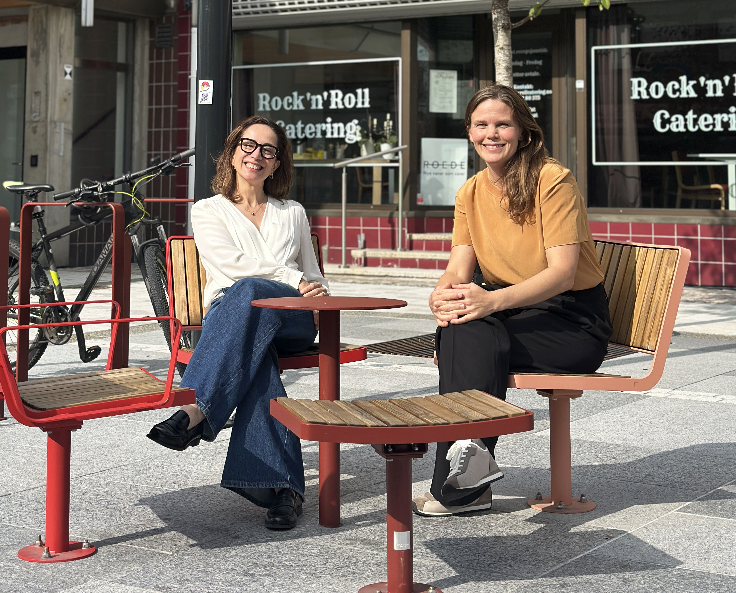 To smilende kvinner sitter utendørs ved siden av et sykkelstativ. Foto.