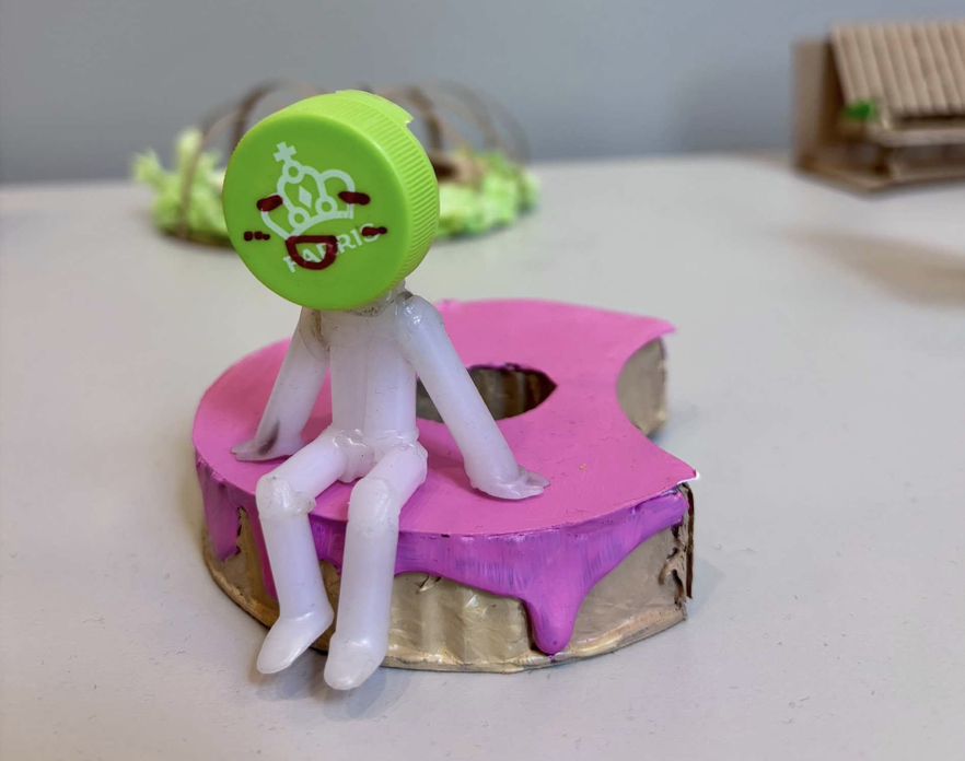 Modell av rosa, rund benk med figur på. Foto.
