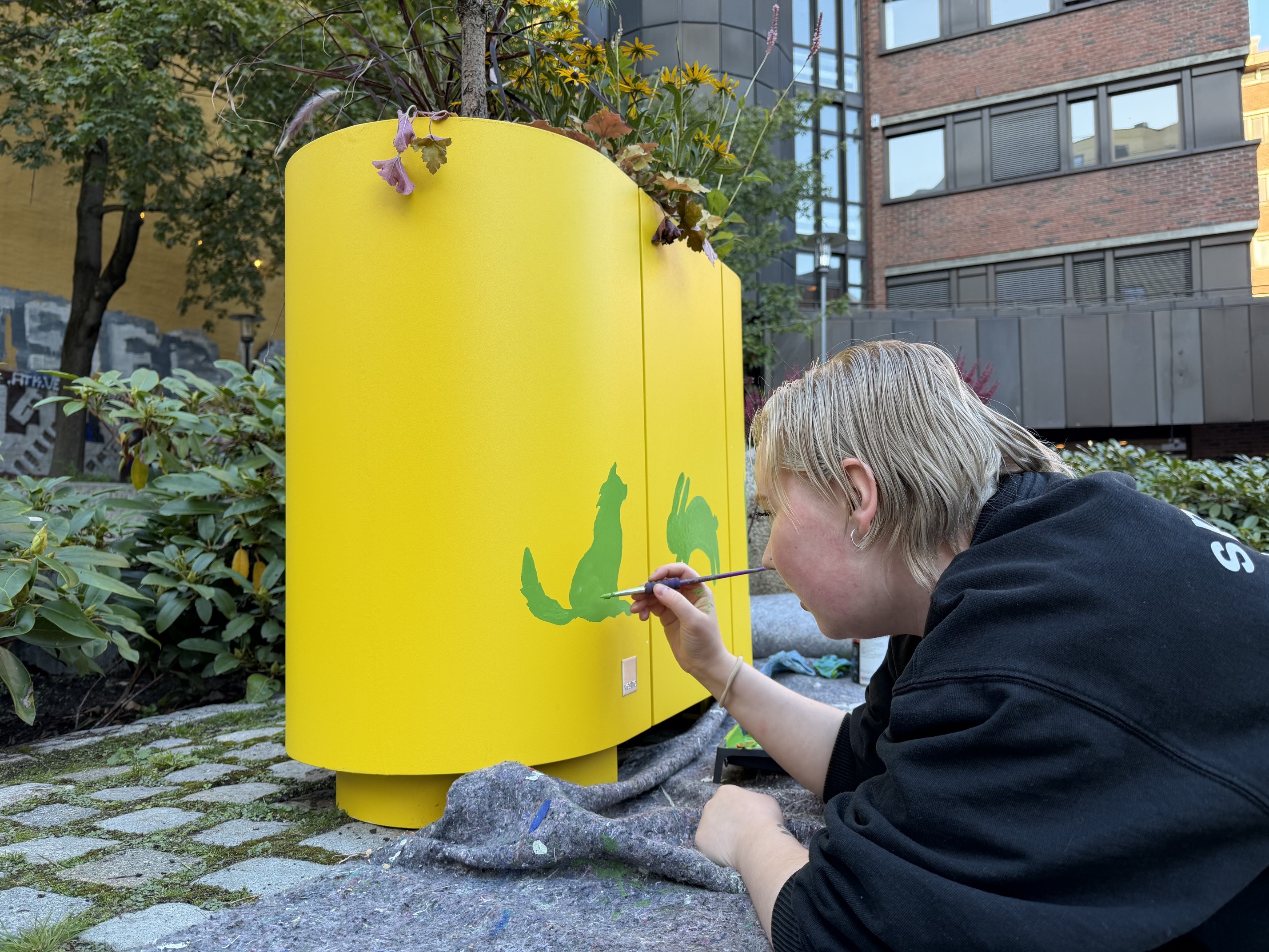 Kvinne maler grønne dyr på gul plantekasse. Foto.